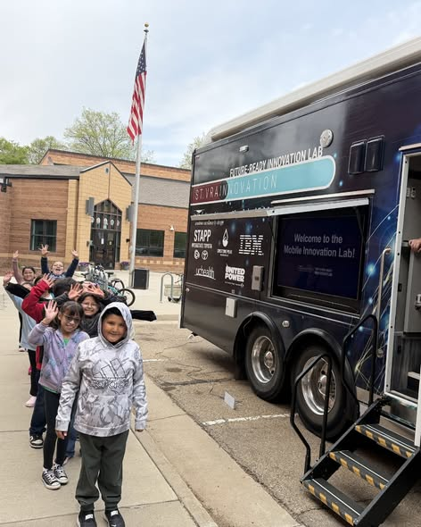 A group of children waves in front of a mobile innovation lab parked near a school, with an American flag in the background.
