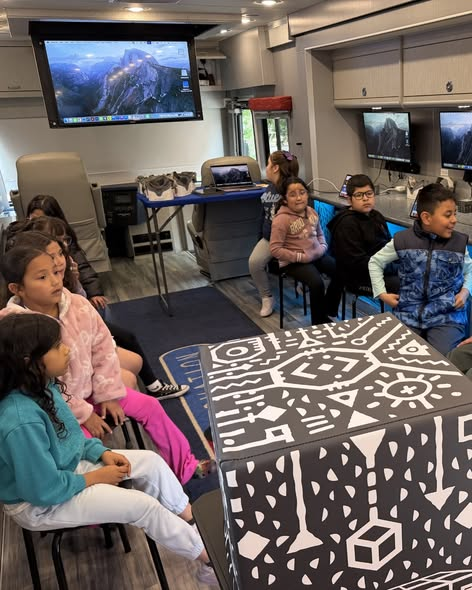 Children are seated in a mobile classroom, engaged, with computers and a large screen displaying a scenic image.