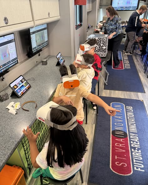 Children wearing virtual reality headsets engage with technology in a classroom setting, with monitors displaying content.