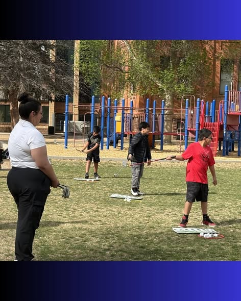 Un grupo de niños practica diversas habilidades en un campo de césped mientras un adulto los supervisa desde cerca, con los juegos infantiles al fondo.
