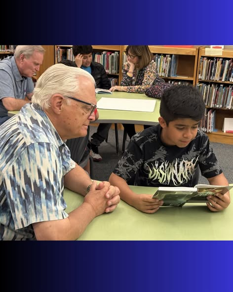 Un hombre mayor y un niño pequeño leen juntos en una biblioteca, mientras que otras personas leen cerca de ellos.