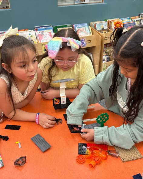 Tres niños están construyendo con bloques de colores sobre una mesa, rodeados de libros en lo que parece ser una biblioteca.