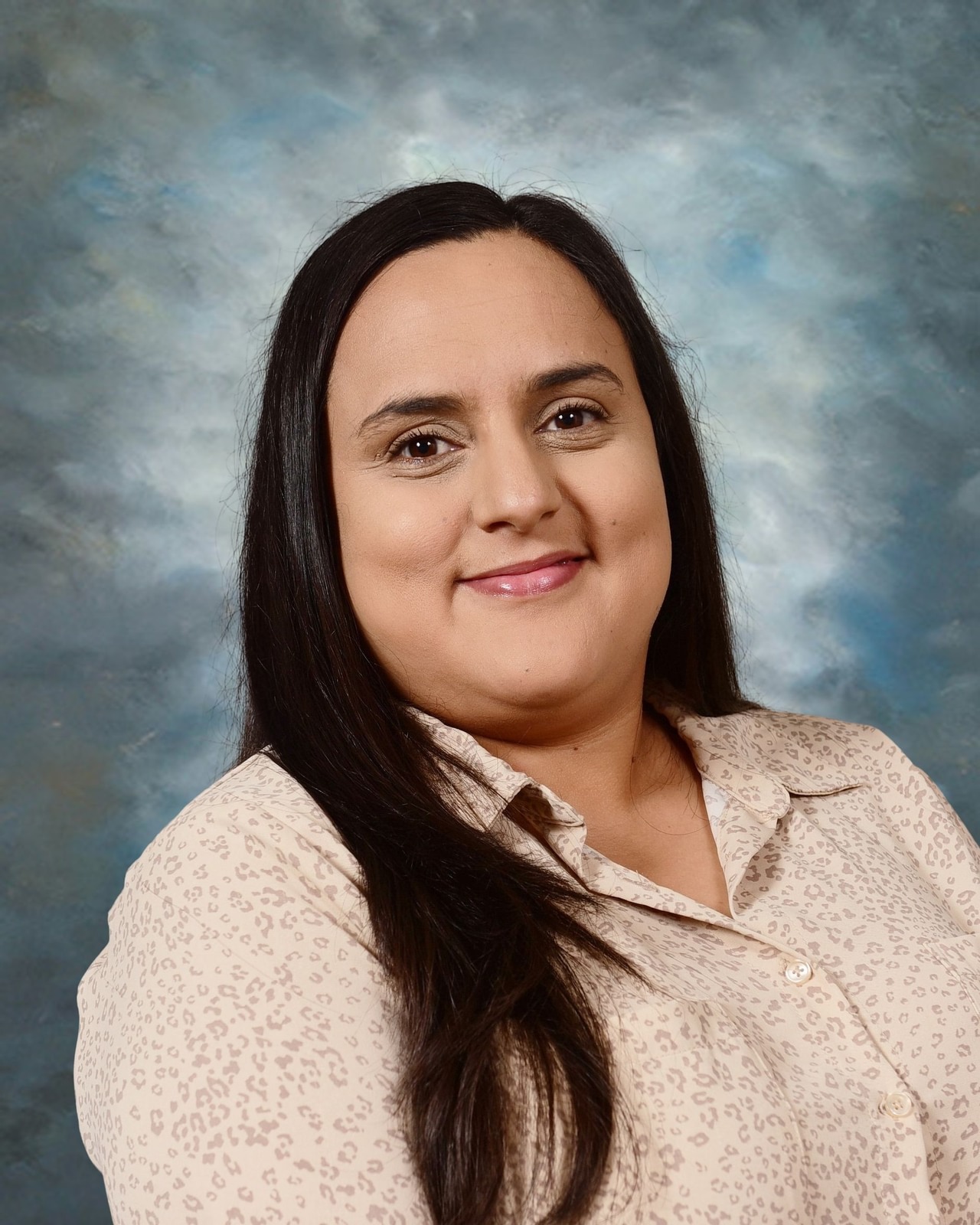 Woman with long dark hair in a light patterned blouse, smiling against a cloudy blue background.