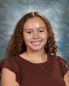 Smiling person with curly hair wearing a brown top, set against a cloudy blue background.