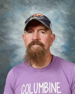 Man with a beard wearing a purple shirt and a cap with a colorful logo, standing against a cloudy background.