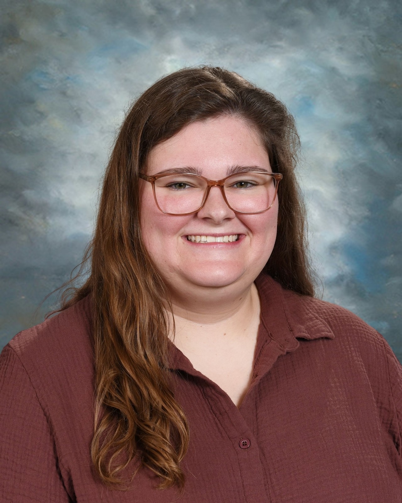 A person with long brown hair and glasses smiles against a cloudy blue background, wearing a brown shirt.