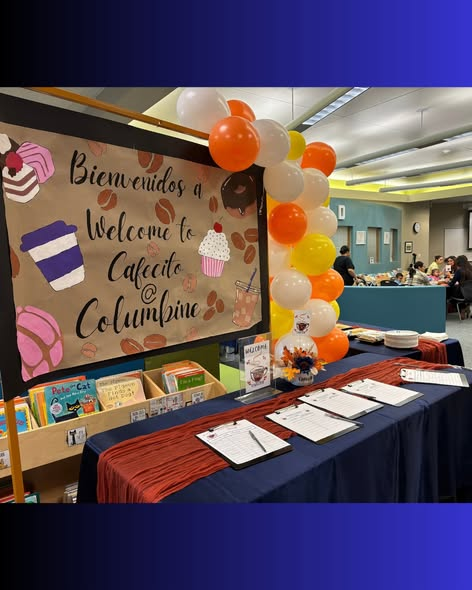 Welcome sign for Cafecito at Columbine, with balloons and a table displaying clipboards, in a room with books and people.
