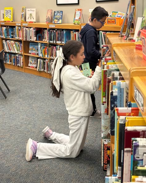 Girl kneeling and boy standing while selecting books from shelves in a library.