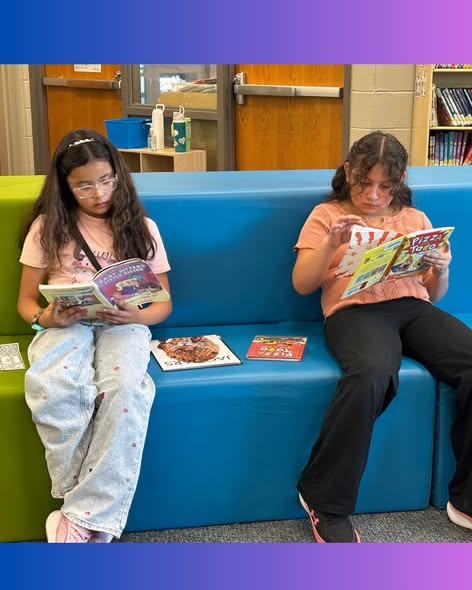 Two girls sit on a colorful library bench, reading comic books. More books are placed on the bench between them.