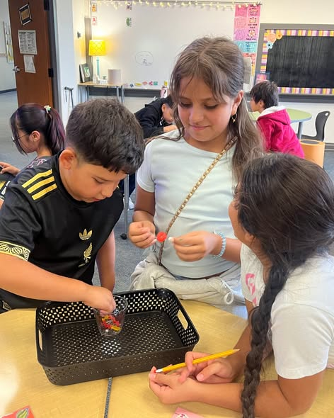 Three children work together at a classroom table, using a black tray and colorful objects.