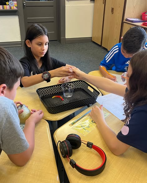 Four children work together at a classroom table, engaging with a small experiment involving a glass and tray.