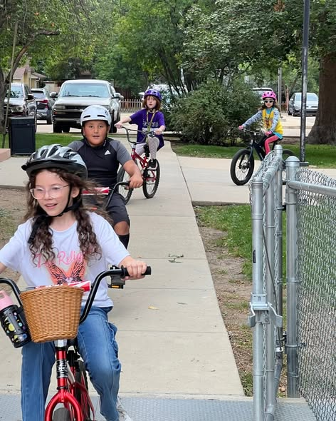 Four children riding bikes on a sidewalk, wearing helmets. Trees and parked cars are in the background.
