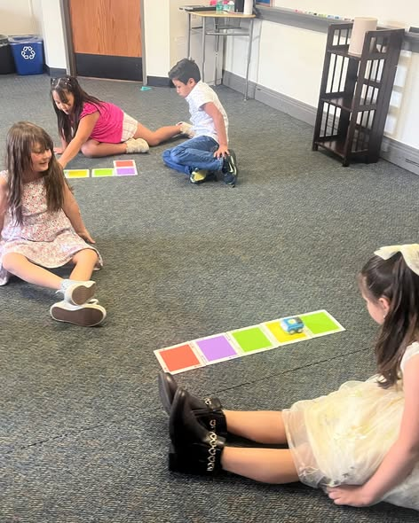 Children sitting on the floor, playing with small robots on colorful mats in a classroom setting.